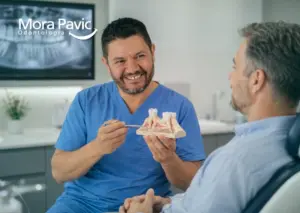 Odontólogo explica a un paciente adulto el procedimiento de injerto óseo dental utilizando un modelo anatómico en la clínica.